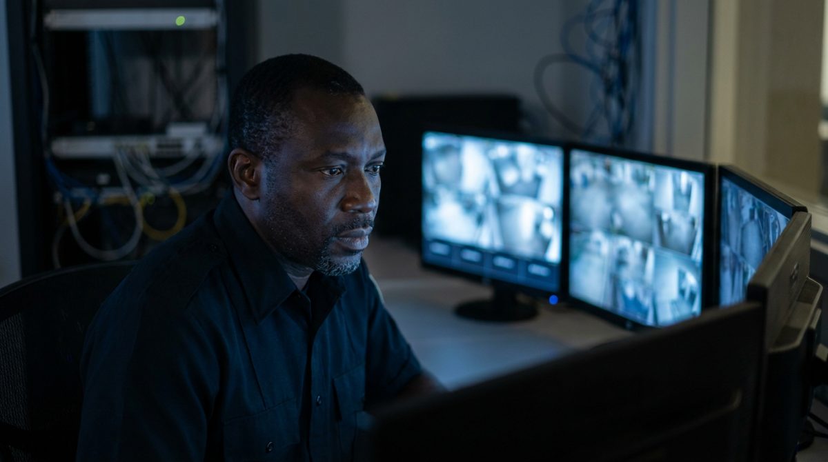 Nigerian security guard monitoring CCTV screens in a Canadian control room at night